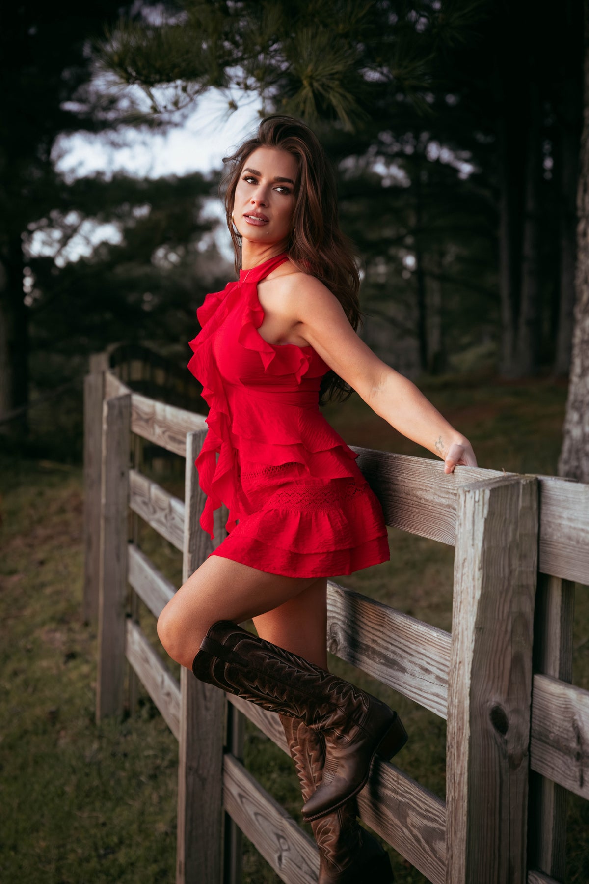 Jessie in a red skirt and top and brown boots leaning against a wooden fence