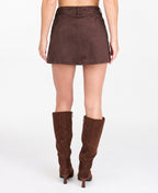 A model wearing a brown suede mini skirt with button and scallop details and matching brown knee-high boots, shown from the waist down on a white studio background.
