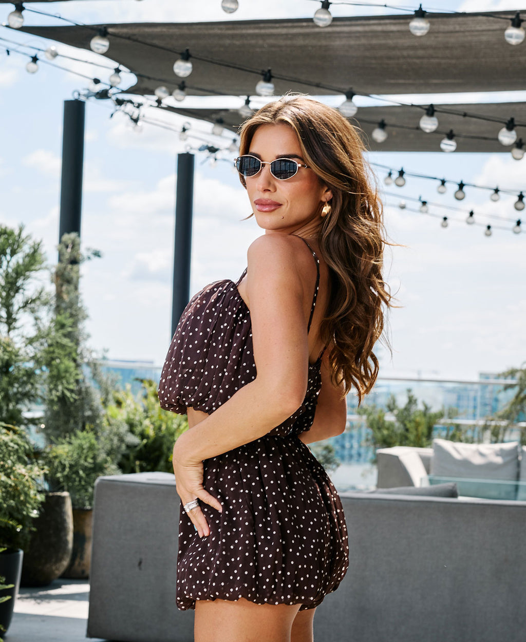 A woman wearing a dark brown and white polka-dot puff-sleeve set and round sunglasses, posing on a rooftop terrace with string lights and a city view in the background.