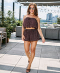 A woman modeling a dark brown two-piece set with white polka dots, consisting of a ruffled spaghetti-strap crop top and matching shorts, on a rooftop terrace.