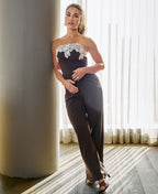 A woman models a dark brown strapless jumpsuit with a white lace bodice detail, standing in a brightly lit room with a large column and sheer curtains.