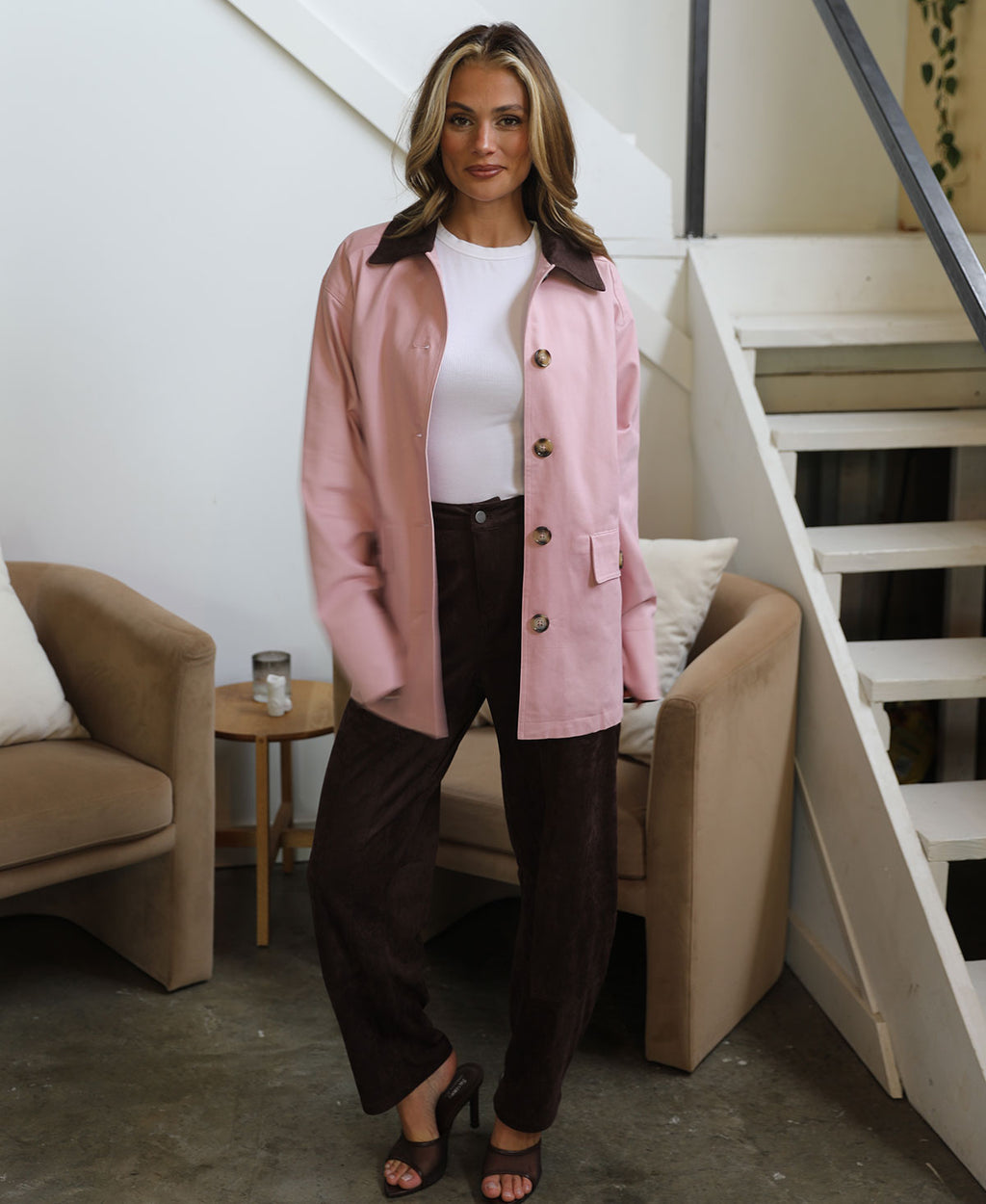 A woman stands indoors wearing a light pink button-front jacket with dark brown trim over a white top and dark brown pants, in a modern room with beige armchairs and a staircase.