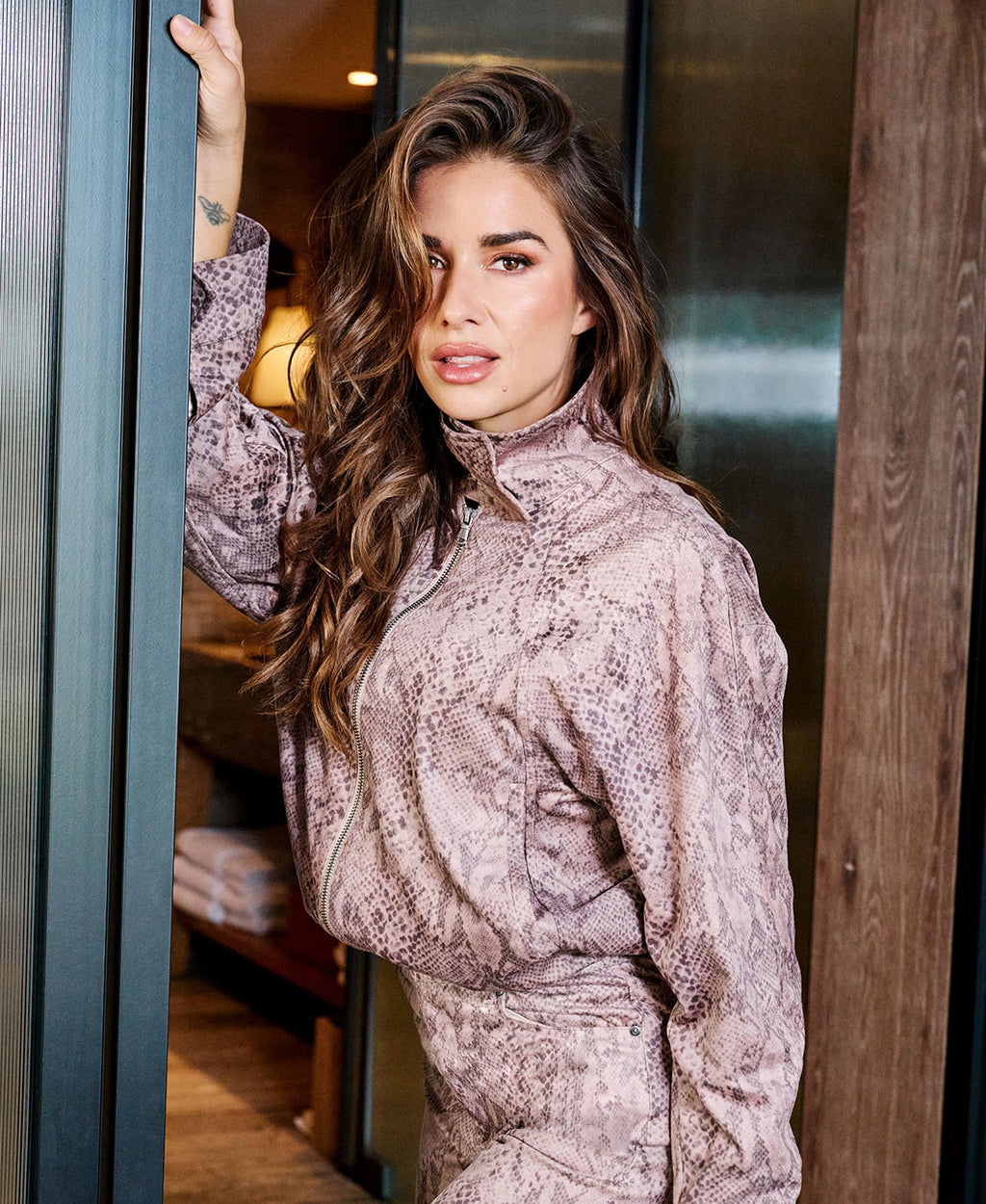 A woman with long wavy brown hair wearing a snakeskin-patterned mauve and brown zip-up jacket, leaning against a dark metal door frame in an indoor setting.