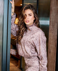 A woman with long wavy brown hair wearing a snakeskin-patterned mauve and brown zip-up jacket, leaning against a dark metal door frame in an indoor setting.
