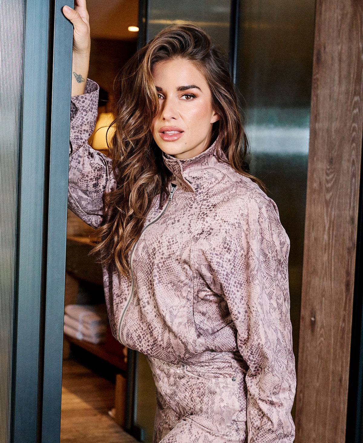 A woman with long wavy brown hair wearing a snakeskin-patterned mauve and brown zip-up jacket, leaning against a dark metal door frame in an indoor setting.