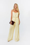 Woman wearing a light yellow jumpsuit holding a brown clutch against a white background