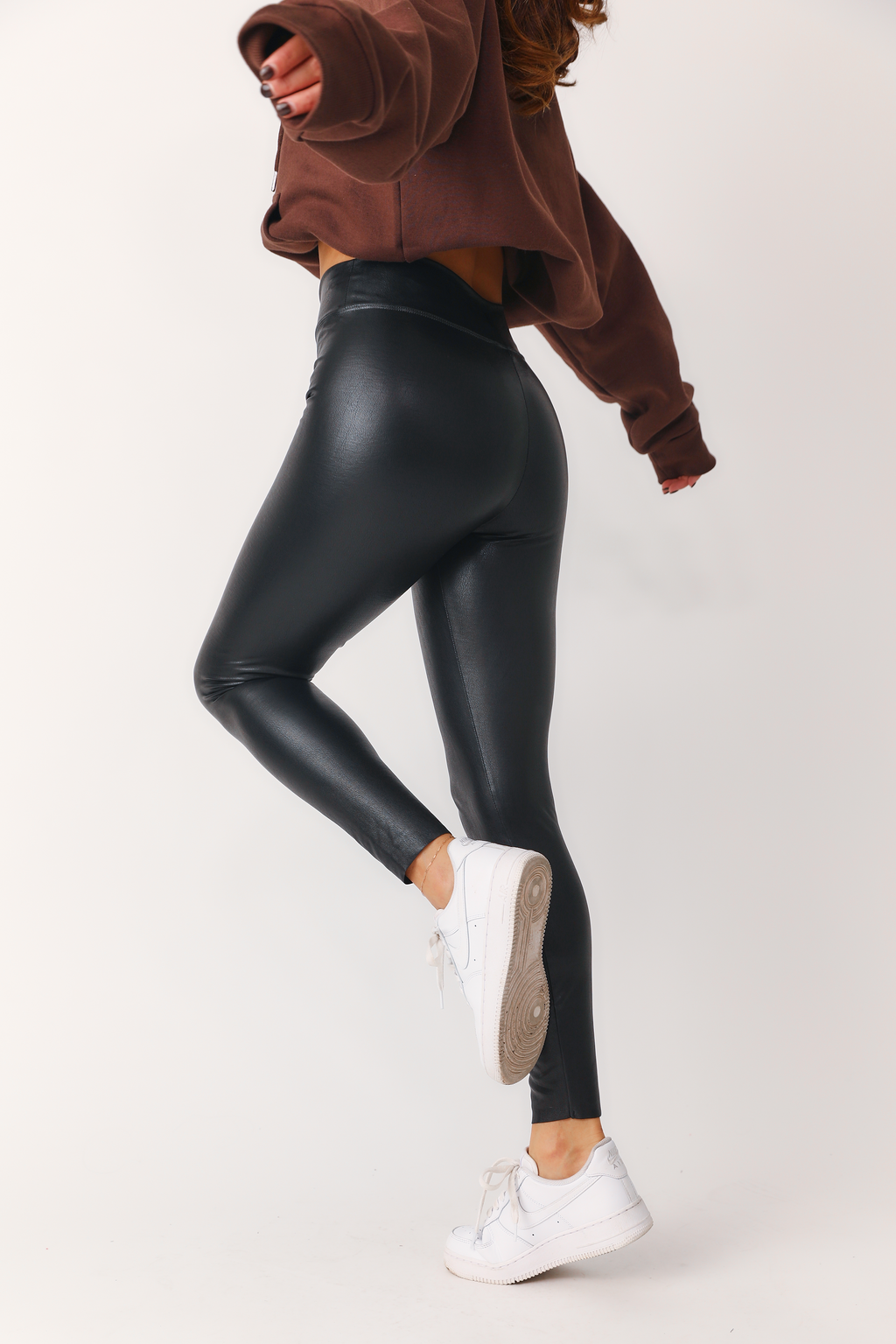 Side/back view of model wearing 'Bet On Me' black faux leather leggings with 'Chocolate Kittenish Logo' hoodie and white sneakers. 