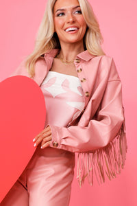 Close up image of model on pink backdrop holding red hearts and modeling the pink Tatum Mae faux leather fringe jacket with matching pink Tatum Mae Faux leather bustier, and pink Tatum Mae faux leather pants. 